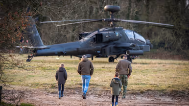 Vier personen, onder wie twee kinderen, lopen naar een militaire helikopter die op een grasveld tussen bomen staat geparkeerd. Rechts: Een militair in vliegtenue met helm en rugzak loopt weg van een grote, grijze militaire helikopter, omringd door bomen en gras. Op de achtergrond zijn kinderen te zien. 
