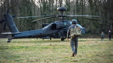 Vier personen, waaronder twee kinderen, lopen naar een militaire helikopter die op een grasveld tussen bomen staat geparkeerd. Rechts: Een militair in vliegtenue met helm en rugzak loopt weg van een grote, grijze militaire helikopter, omringd door bomen en gras. Op de achtergrond zijn kinderen te zien. 