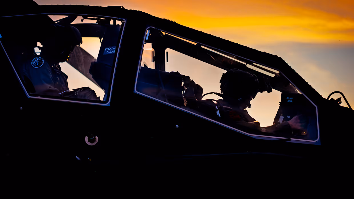 Een zijdelingse close-up van een Apache cockpit met twee piloten erin die van achter belicht worden door schemerend zonlicht.