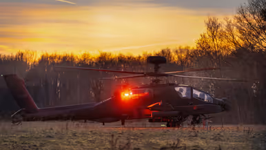 Een militaire helikopter staat stil op een grasveld tijdens zonsondergang, met rode navigatielichten aan en bomen op de achtergrond. Rechts: Een silhouet van een militaire helikopter vliegt laag tijdens zonsondergang met draaiende rotorbladen tegen een gekleurde lucht met bomen op de achtergrond.