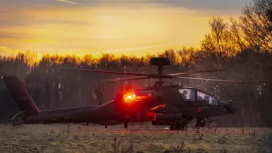 Een militaire helikopter staat stil op een grasveld tijdens zonsondergang, met rode navigatielichten aan en bomen op de achtergrond. Rechts: Een silhouet van een militaire helikopter vliegt laag tijdens zonsondergang met draaiende rotorbladen tegen een gekleurde lucht met bomen op de achtergrond.