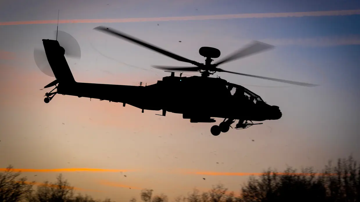 Een militaire helikopter staat stil op een grasveld tijdens zonsondergang, met rode navigatielichten aan en bomen op de achtergrond. Rechts: Een silhouet van een militaire helikopter vliegt laag tijdens zonsondergang met draaiende rotorbladen tegen een gekleurde lucht met bomen op de achtergrond.