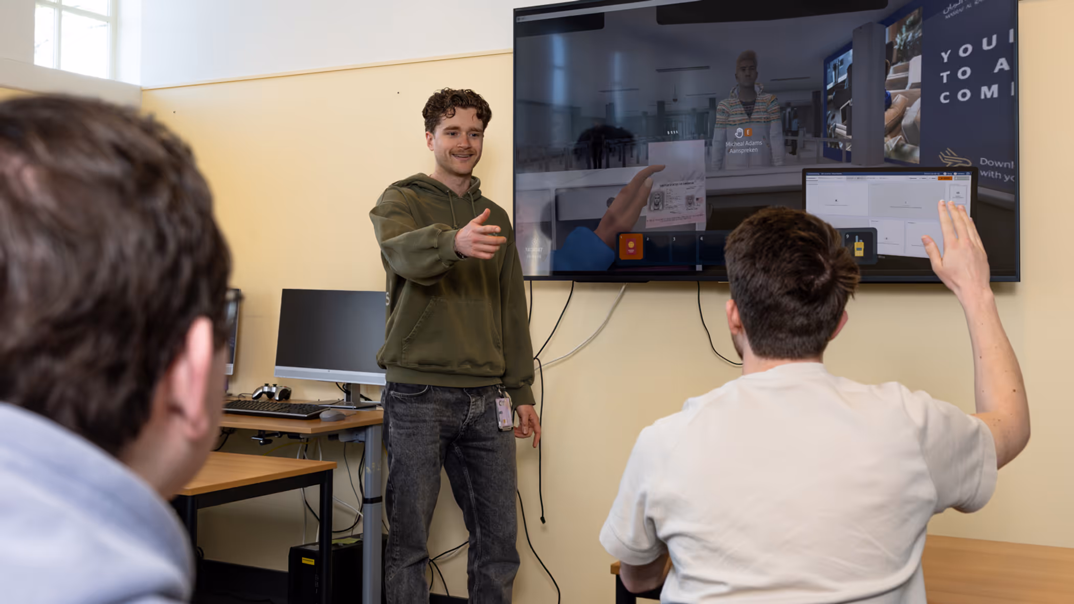 Een begeleider in een groene trui staat voor een digitaal schoolbord. Hij geeft de beurt aan een stagiair die zijn hand opsteekt.