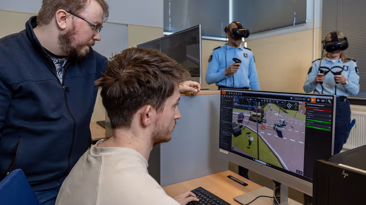 Twee mannen kijken naar een computerscherm waarop een animatie van een verkeerssituatie is te zien. Op de achtergrond hebben twee marechaussees een VR-bril op hun hoofd en controllers in hun handen.