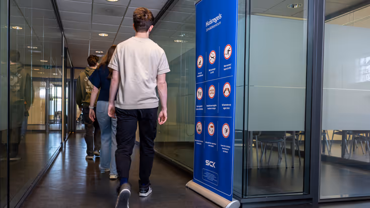  Jongeren lopen door een gang (op de rug gezien). Ze passeren een manshoog, blauw bord met allerlei icoontjes erop. 