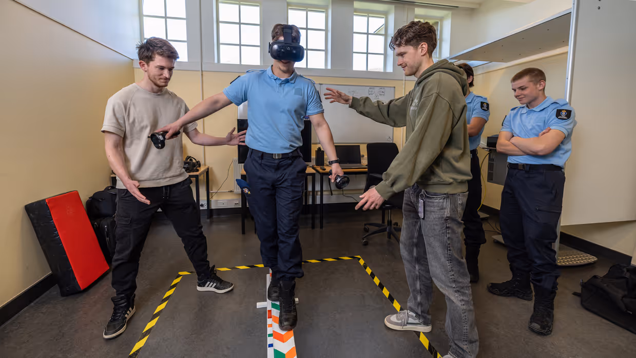 Een marechaussee in opleiding draagt een VR-bril en balanceert op een balk. Twee burgercollega’s begeleiden hem, terwijl op de achtergrond twee marechaussees toekijken.