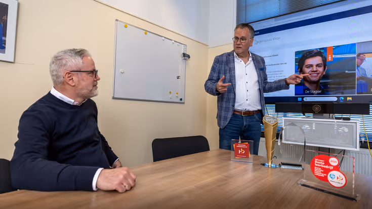 Een man staat bij een televisiescherm en wijst naar een foto van een stagiair. Links van hem zit een man te luisteren naar de uitleg. 