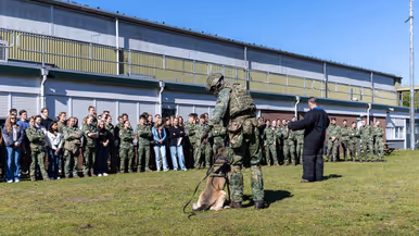  Jonge militairen en enkele burgers vormen een halve cirkel op het gras rondom een militair met een hond en een pakwerker die uitleg geeft.