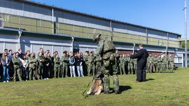  Jonge militairen en enkele burgers vormen een halve cirkel op het gras rondom een militair met een hond en een pakwerker die uitleg geeft.