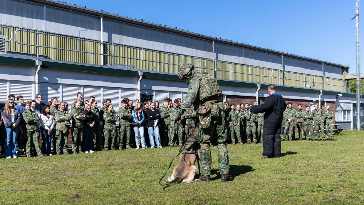  Jonge militairen en enkele burgers vormen een halve cirkel op het gras rondom een militair met een hond en een pakwerker die uitleg geeft.