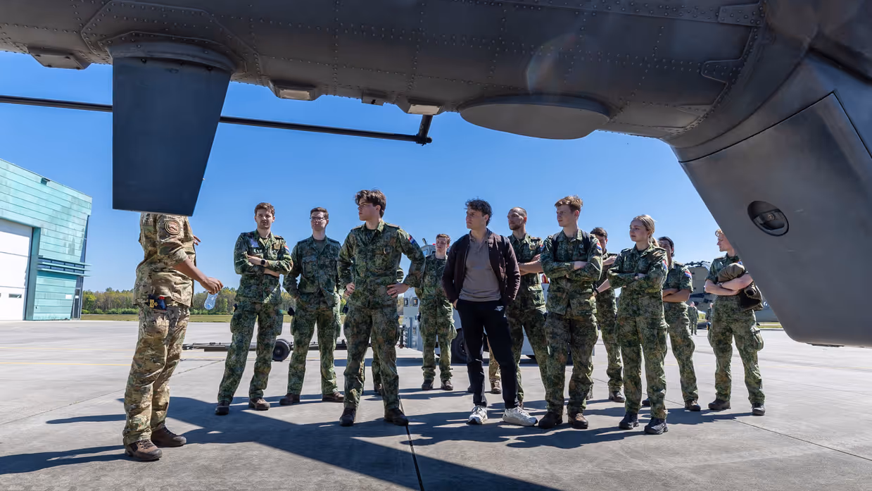 Jonge militairen en een burger krijgen uitleg van een militair   naast een helikopter. 