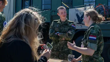 Een militair van het MAOT staand achter een tafel in gesprek met een jonge militair met een sticker in de hand. Rondom de tafel staan meerdere burgers en militairen.