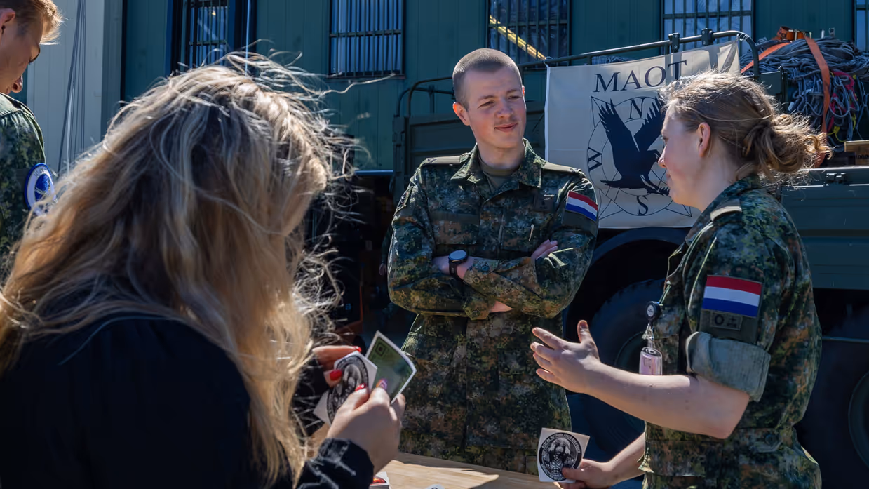 Een militair van het MAOT staand achter een tafel in gesprek met een jonge militair met een sticker in de hand. Rondom de tafel staan meerdere burgers en militairen.