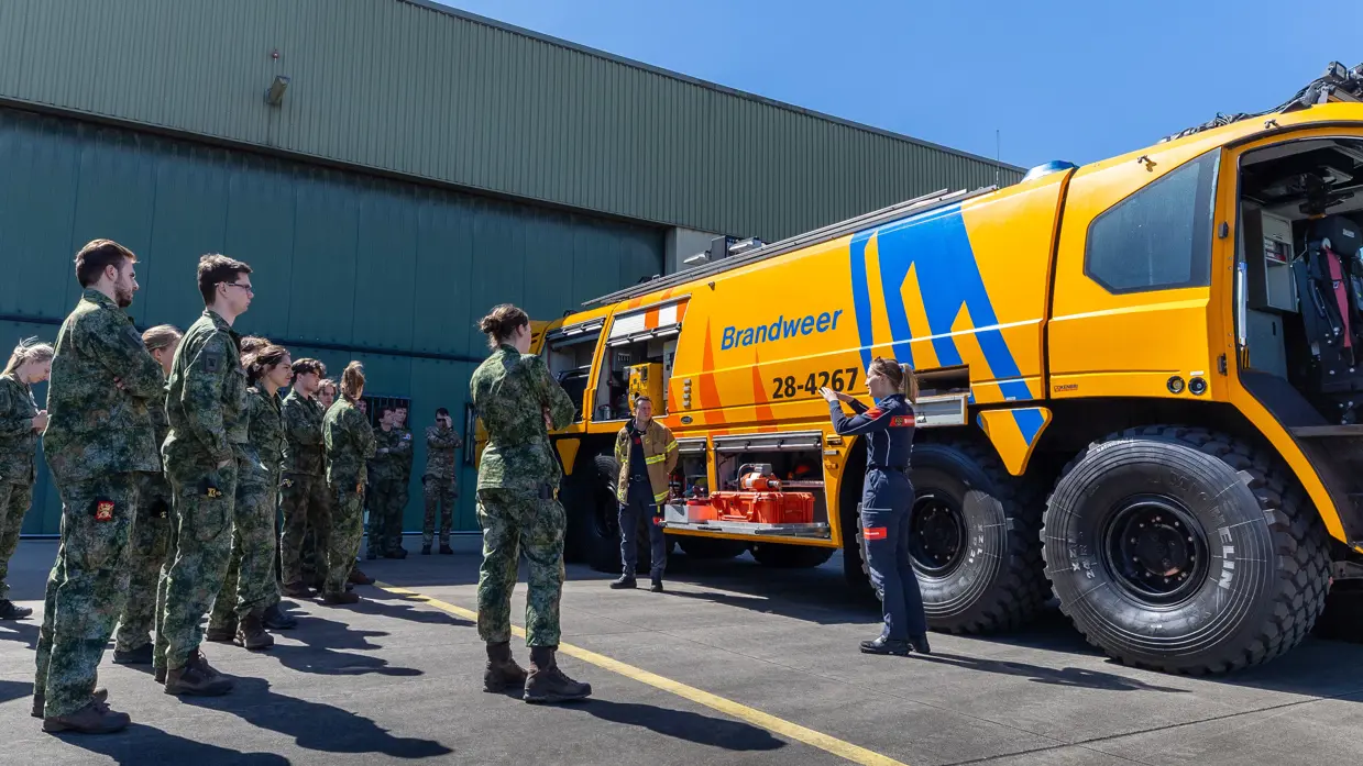 Een militair van de brandweer naast een gele brandweerwagen geeft uitleg aan een groepje jonge militairen.
