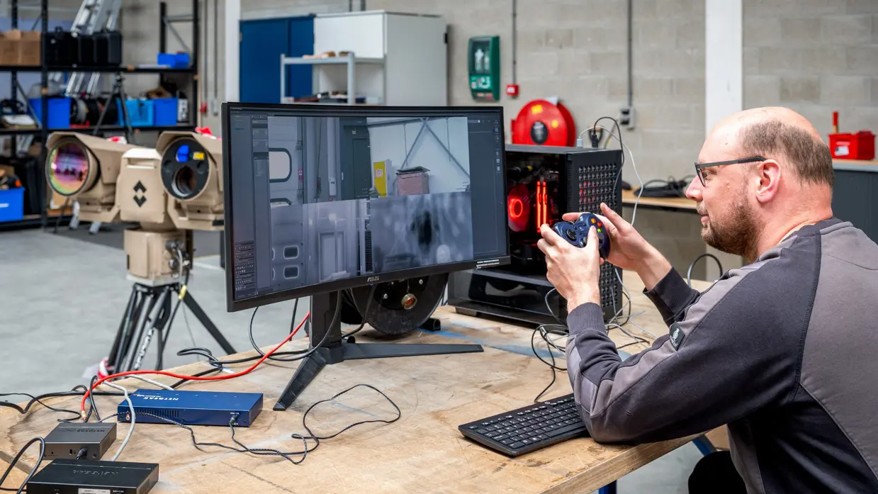 Een man heeft een soort joystick in zijn hand en kijkt naar een beeldscherm op de tafel voor hem. 