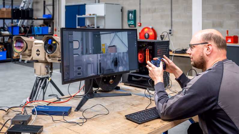 Een man heeft een soort joystick in zijn hand en kijkt naar een beeldscherm op de tafel voor hem. 