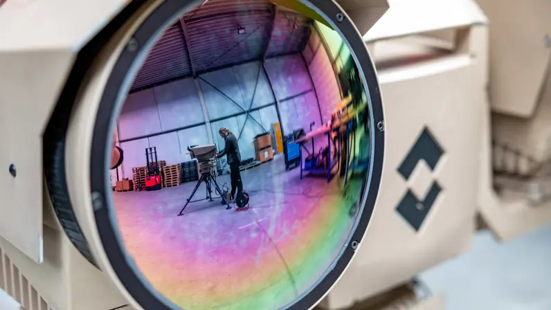 Een foto van een grote cameralens, waarin je in de weerspiegeling iemand op de achtergrond ziet staan.