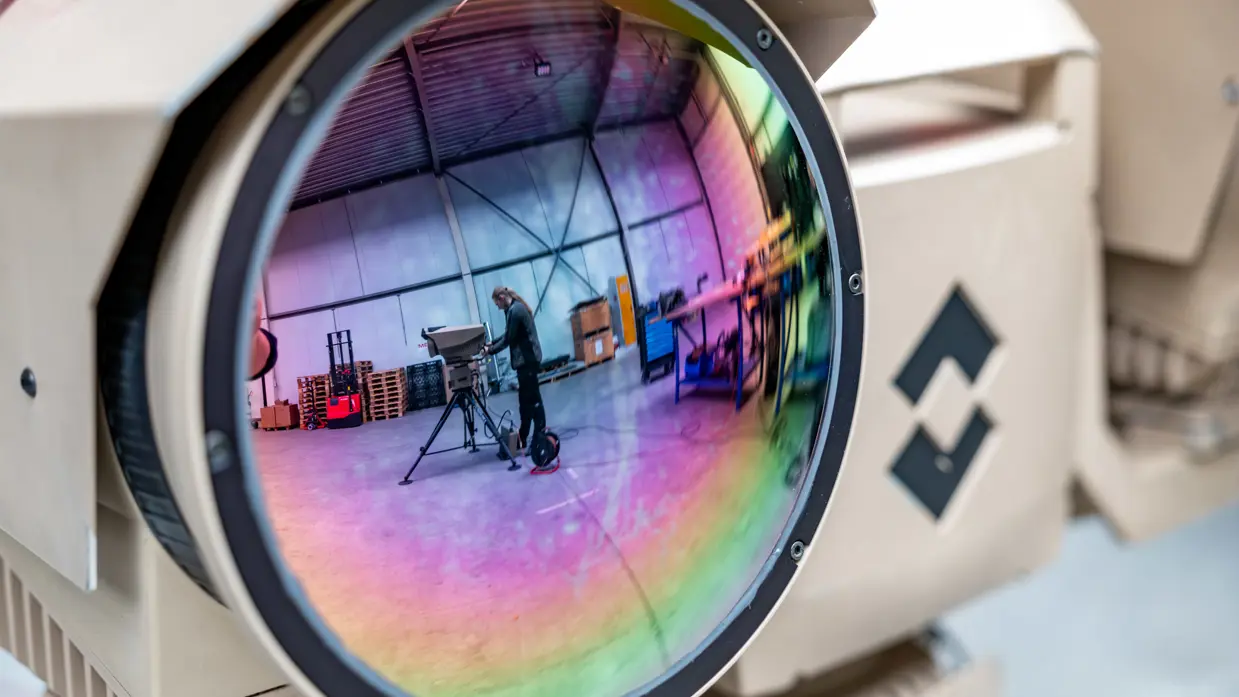 Een foto van een grote cameralens, waarin je in de weerspiegeling iemand op de achtergrond ziet staan.