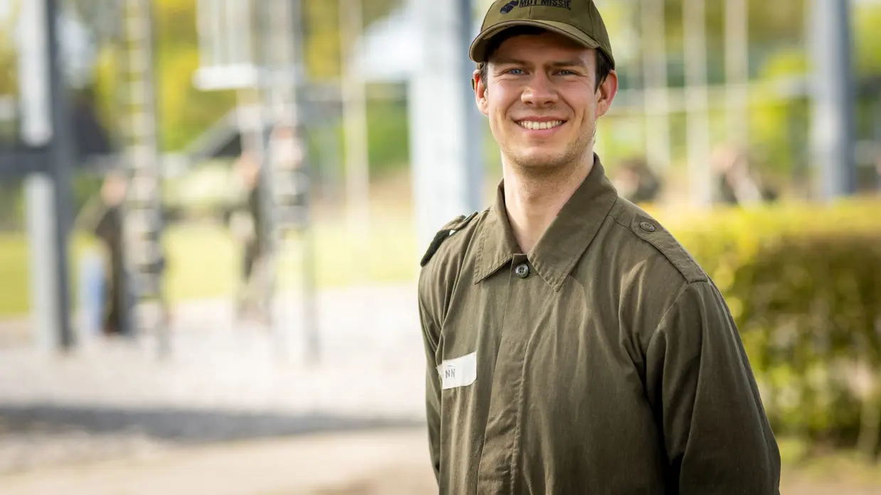MDT-deelnemer Finn staat glimlachend in uniform en pet op de kazerne, met een oefenparcours op de achtergrond. 