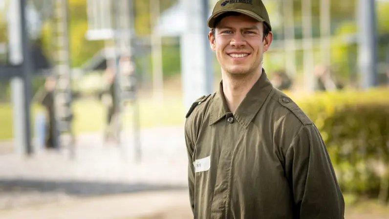MDT-deelnemer Finn staat glimlachend in uniform en pet op de kazerne, met een oefenparcours op de achtergrond. 