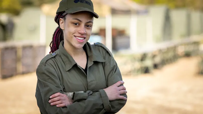 MDT-deelnemer Fiona in militair tenue en pet staat met haar armen over elkaar op de kazerne en kijkt lachend in de camera. 