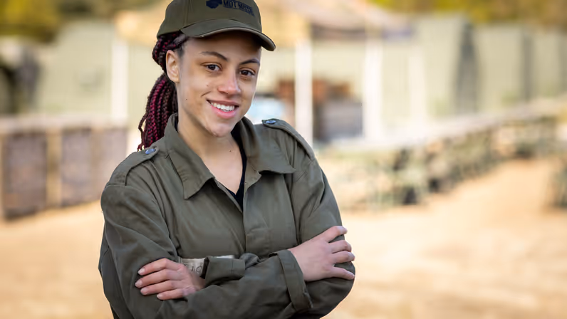 MDT-deelnemer Fiona in militair tenue en pet staat met haar armen over elkaar op de kazerne en kijkt lachend in de camera. 