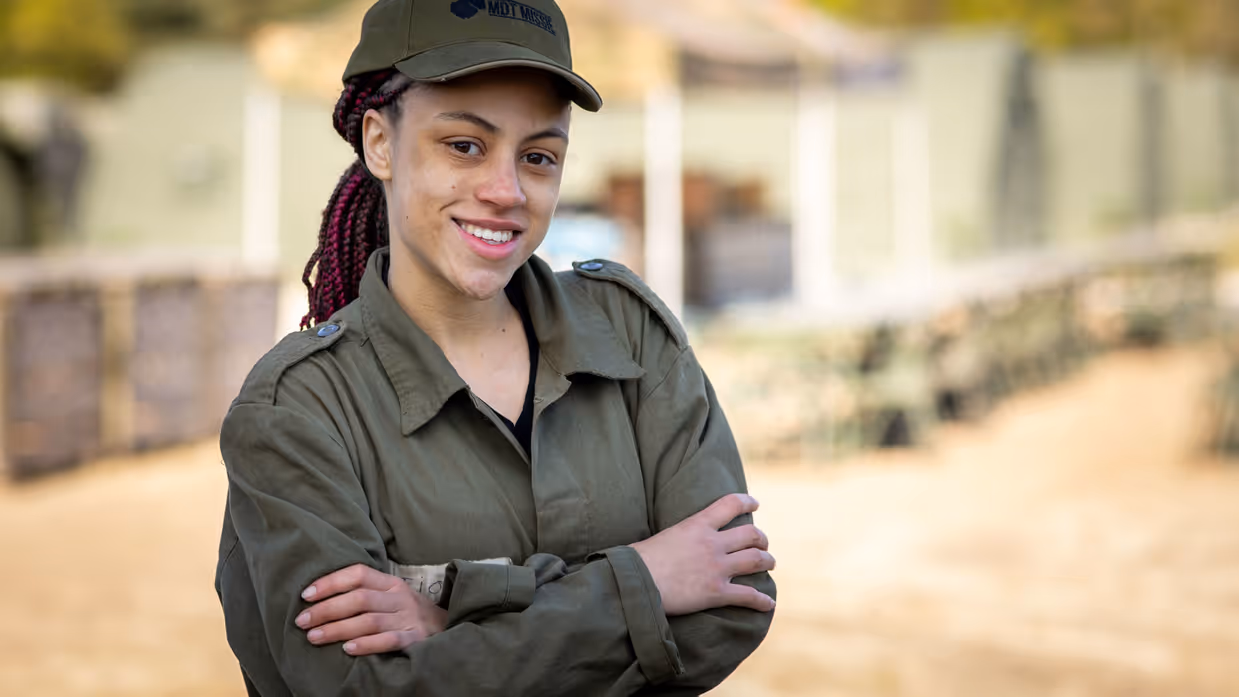 MDT-deelnemer Fiona in militair tenue en pet staat met haar armen over elkaar op de kazerne en kijkt lachend in de camera. 