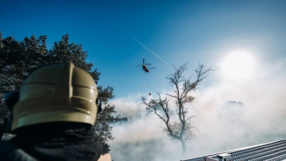 Een brandweerman kijkt vanaf de grond naar een overvliegende helikopter die water loost op een brandhaard. 