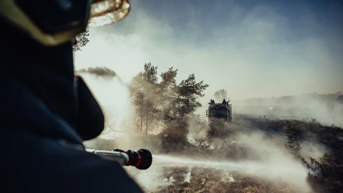 Een brandweerman hanteert een blusapparaat op een voertuig. Op de achtergrond zijn andere collega’s op een brandweerwagen ook bezig met het blussen van de brand. 