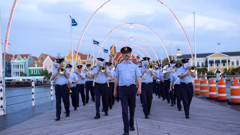 Orkest met blazers vooraan loopt achter man aan brug met lampjes over. 