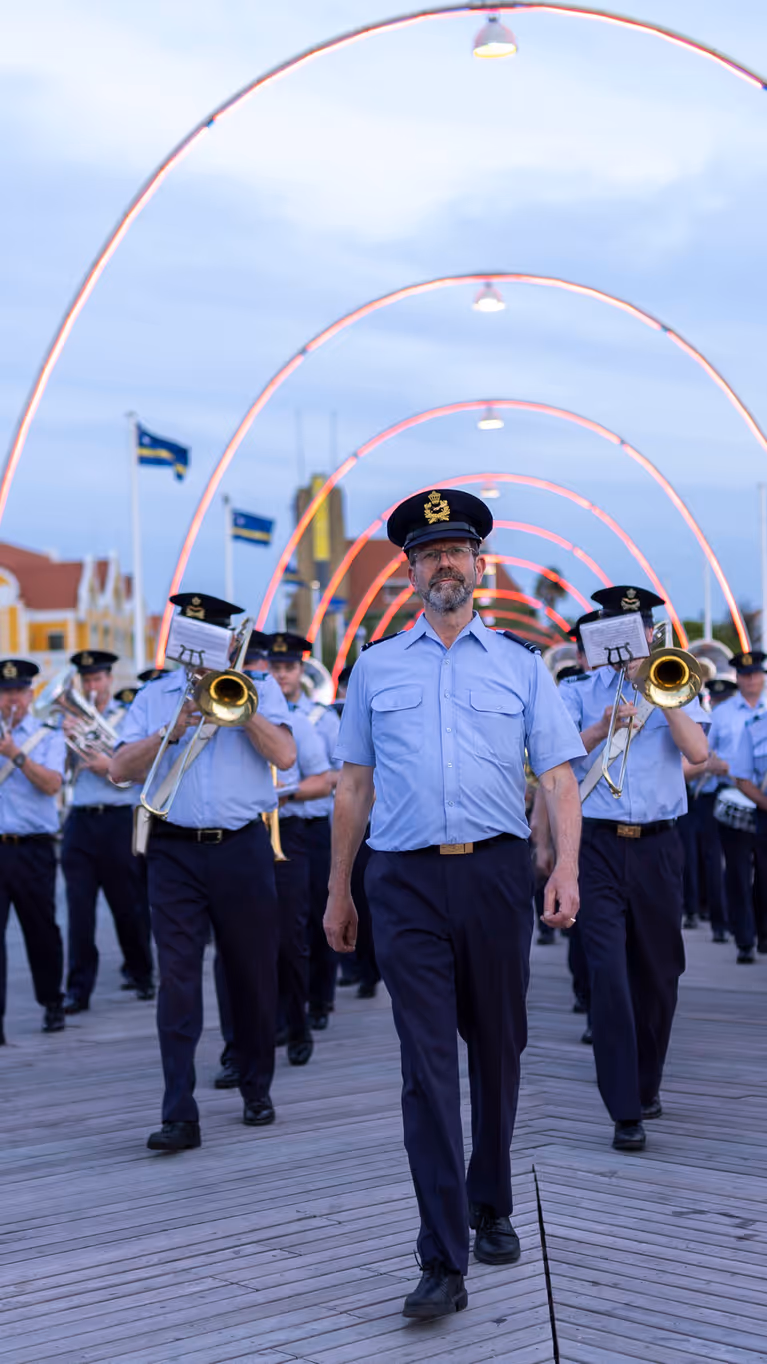 Orkest met blazers vooraan loopt achter man aan brug met lampjes over. 