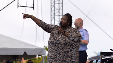 Donkere bebrilde zangeres zingt op podium en wijst naar publiek. 
