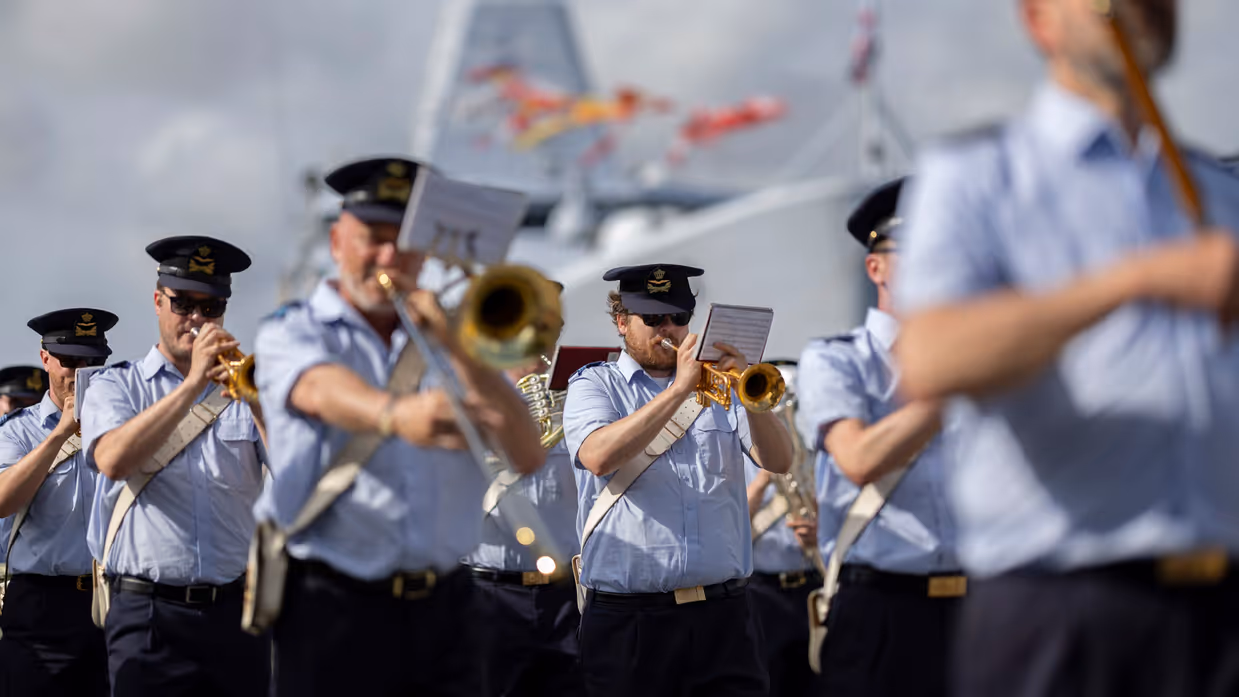 Muzikanten van luchtmacht spelen op hun instrumenten met op achtergrond een marineschip. 