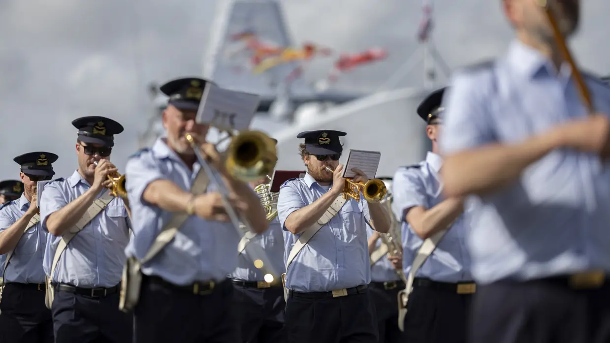 Muzikanten van luchtmacht spelen op hun instrumenten met op achtergrond een marineschip. 