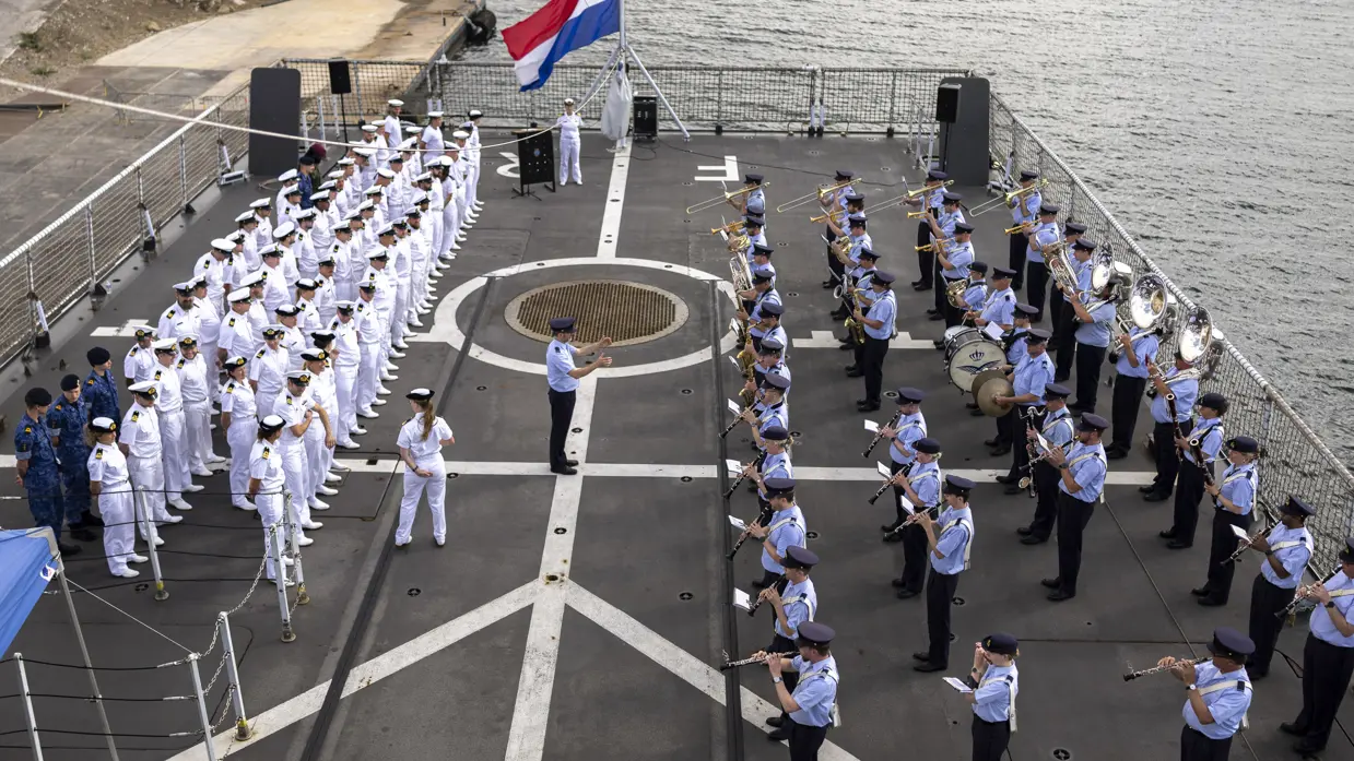 Op het dek van een aangemeerd schip staat rechts het Orkest KLu te spelen tegenover de in rijen opgestelde in wit geklede scheepsbemanning. In het midden tegen de railing hangt de Nederlandse vlag halfstok.