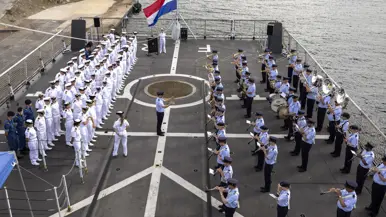 Op het dek van een aangemeerd schip staat rechts het Orkest KLu te spelen tegenover de in rijen opgestelde in wit geklede scheepsbemanning. In het midden tegen de railing hangt de Nederlandse vlag halfstok.