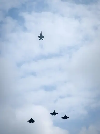 Drie F-35’s vliegen in formatie, terwijl de vierde recht omhoog vliegt naar een half bewolkte lucht. 