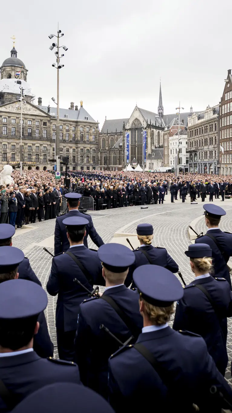 Beeld van de Dam in Amsterdam met de blik pop het Paleis op de Dam, met in de voorgrond op de rug gekeken opgestelde luchtmachtmilitairen. Daartegenover staan vele honderden genodigden en bezoekers van de herdenking.