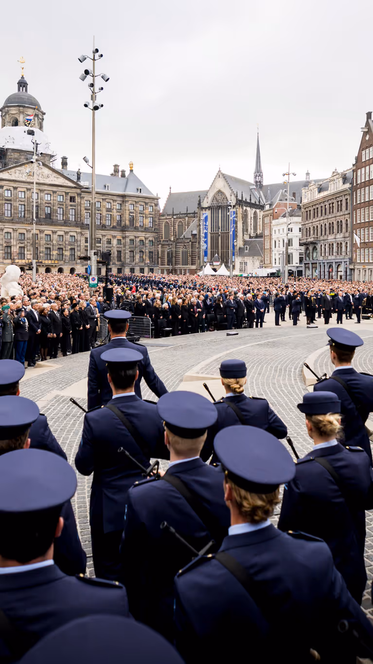 Beeld van de Dam in Amsterdam met de blik pop het Paleis op de Dam, met in de voorgrond op de rug gekeken opgestelde luchtmachtmilitairen. Daartegenover staan vele honderden genodigden en bezoekers van de herdenking.