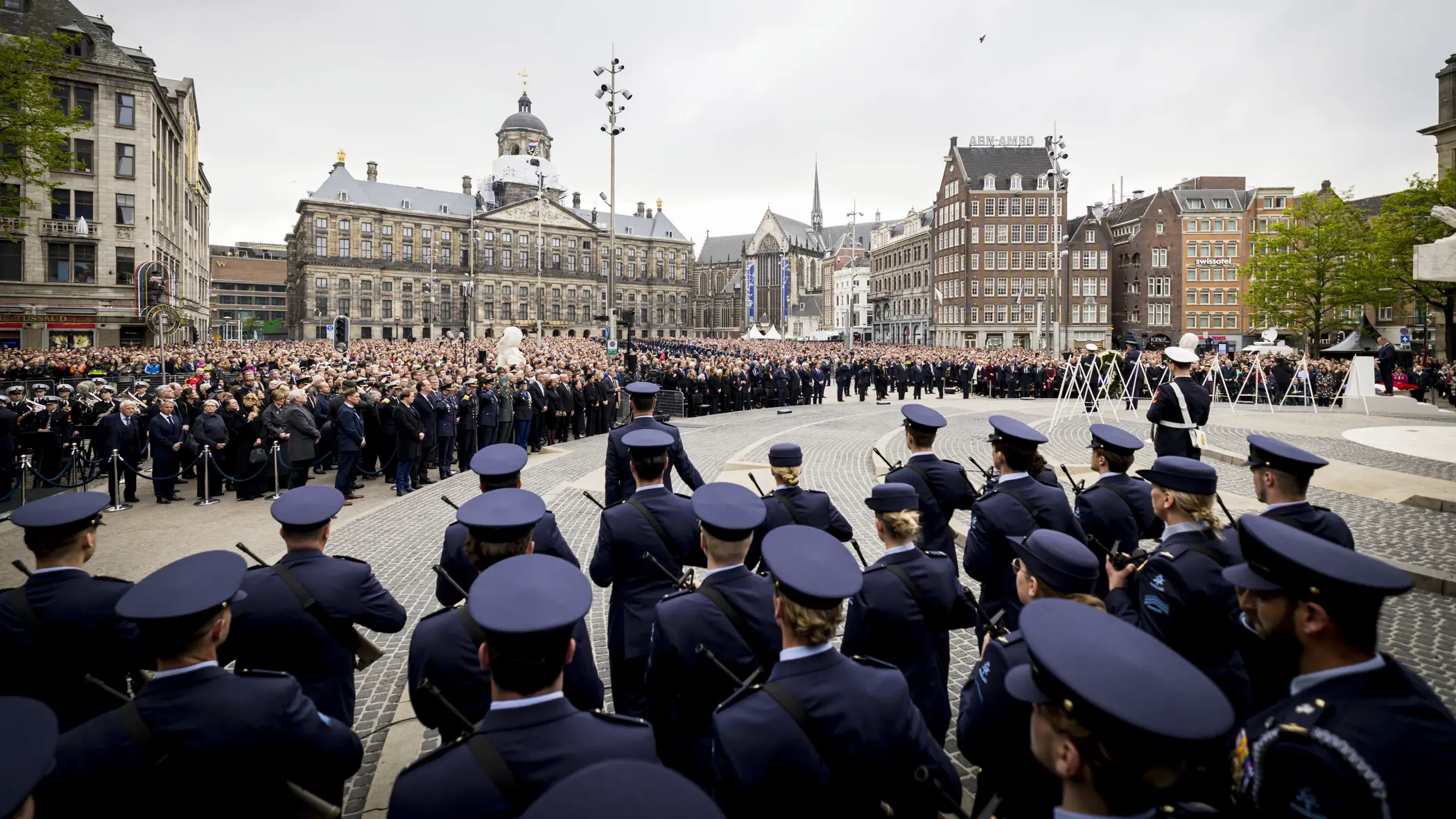 Beeld van de Dam in Amsterdam met de blik pop het Paleis op de Dam, met in de voorgrond op de rug gekeken opgestelde luchtmachtmilitairen. Daartegenover staan vele honderden genodigden en bezoekers van de herdenking.