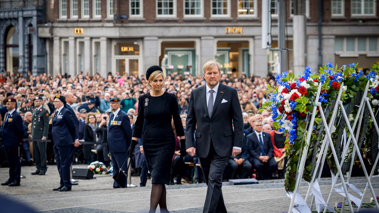 De koning en koningin lopen langs de kransen die rechts te zien zijn. In de achtergrond is publiek te zien.