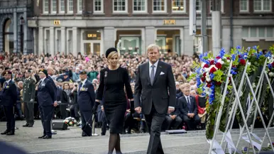 De koning en koningin lopen langs de kransen die rechts te zien zijn. In de achtergrond is publiek te zien.