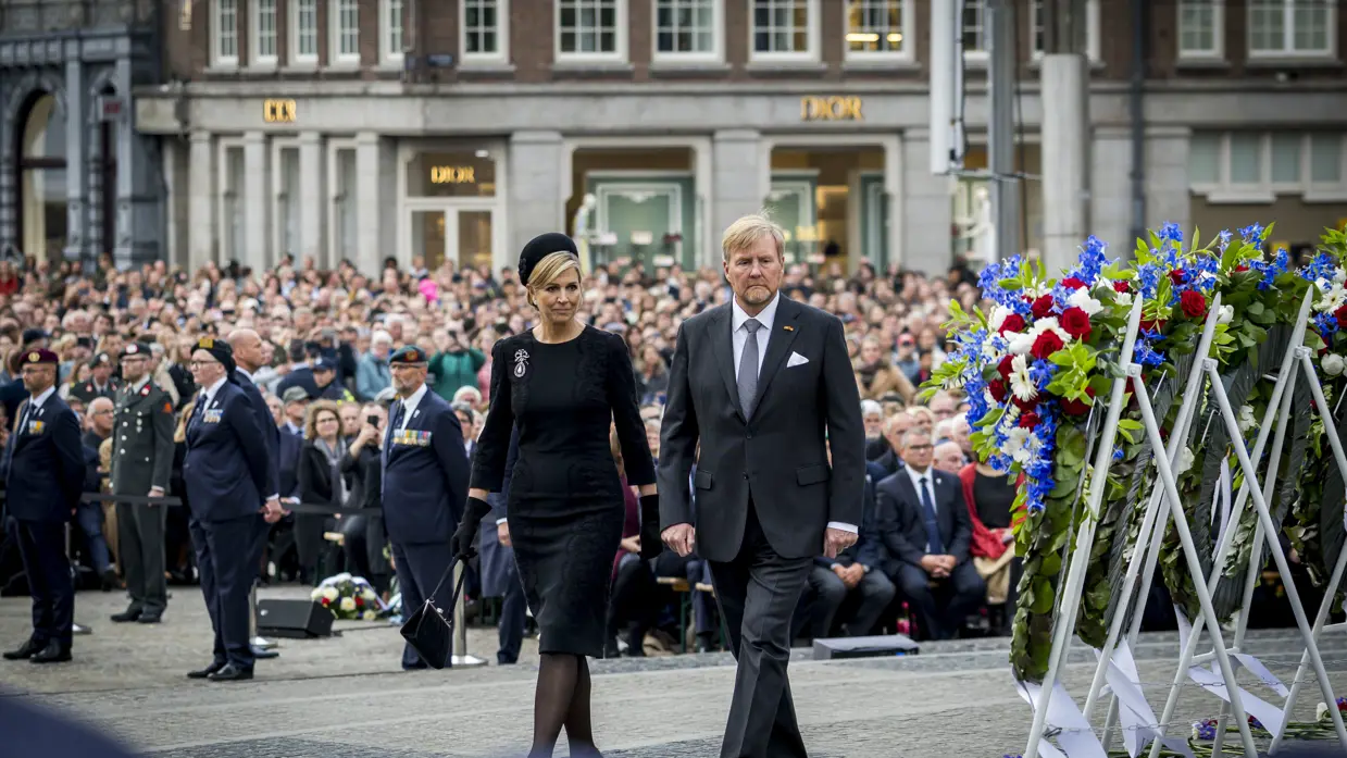 De koning en koningin lopen langs de kransen die rechts te zien zijn. In de achtergrond is publiek te zien.