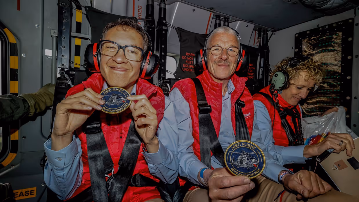 Twee leden van de Jostiband zittend in een NH90 met tonen hun patch ‘Josti Flight; Bevrijdingsdag 2026’.