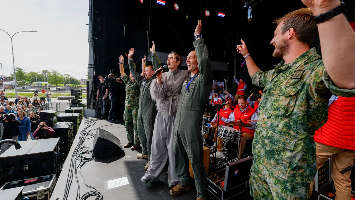  Militairen op een podium juichen met handen in de lucht naar het publiek. Te midden van hen zangeres Maan. Achter hen de Jostiband.