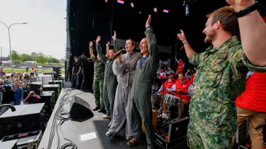  Militairen op een podium juichen met handen in de lucht naar het publiek. Te midden van hen zangeres Maan. Achter hen de Jostiband.