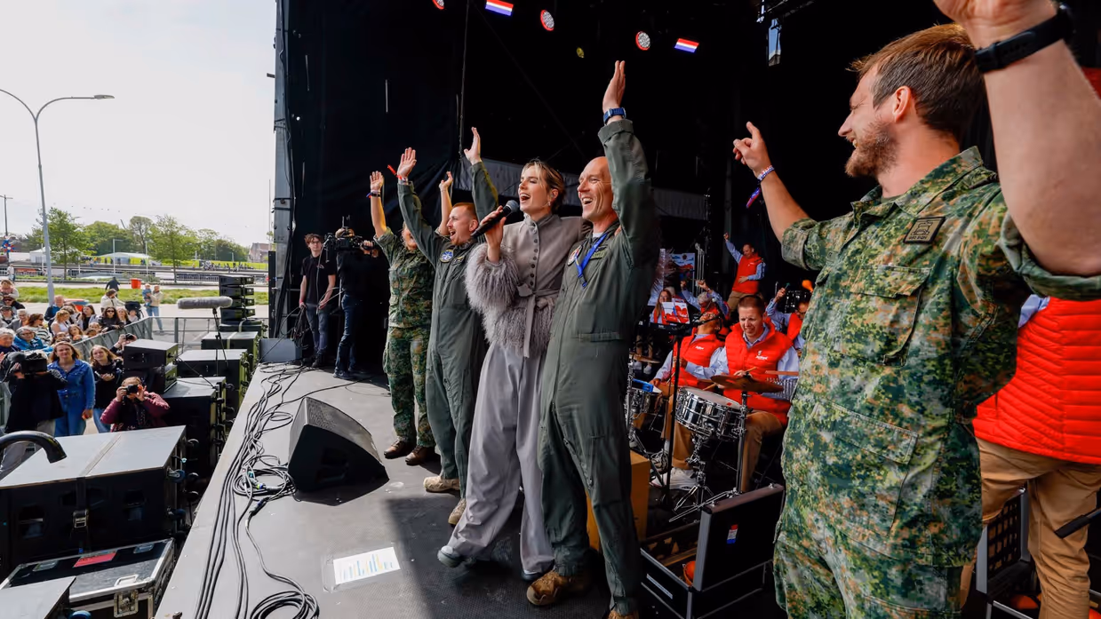  Militairen op een podium juichen met handen in de lucht naar het publiek. Te midden van hen zangeres Maan. Achter hen de Jostiband.