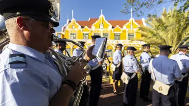 Luchtmachtmuzikanten treden op tegen een achtergrond van gele huizen en palmbomen.