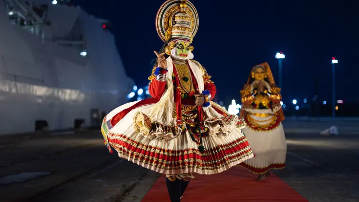 Een traditioneel geklede en gemaskerde Indiër op een rode loper naast de De Ruyter.  &nbsp;