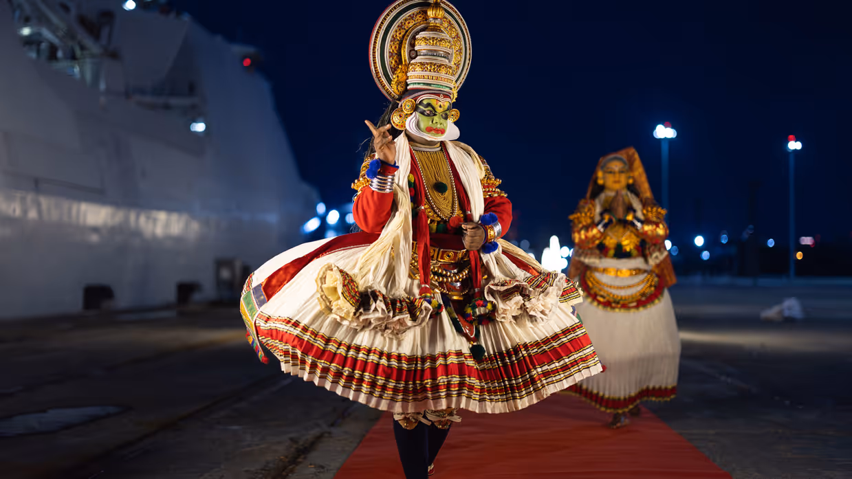 Een traditioneel geklede en gemaskerde Indiër op een rode loper naast de De Ruyter.  &nbsp;